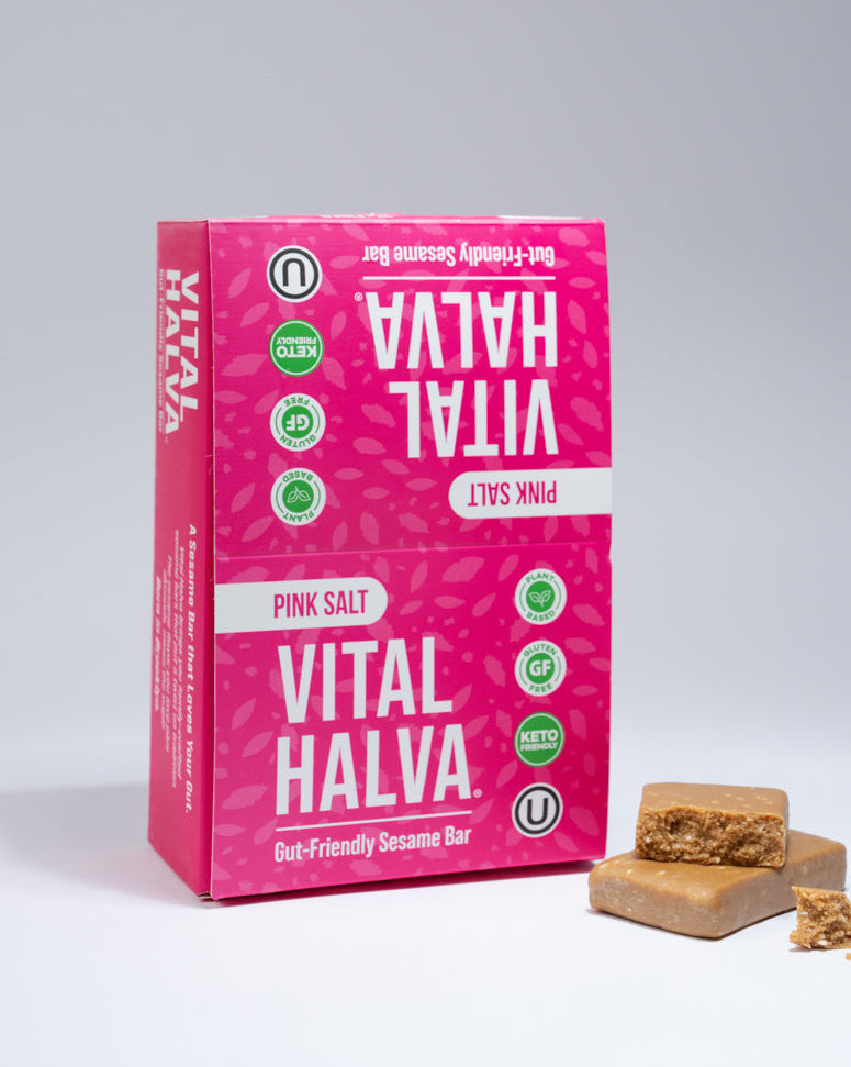 A pink box of Vital Halva Pink Salt sesame snack bars stands on a light gray studio background, with two sesame snack bars and a small pile of crumb placed in front.